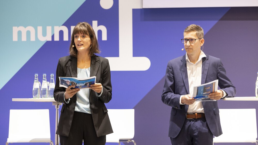 Eine Frau und ein Mann in Anzügen mit bedruckten Seiten in der Hand stehen auf einer Bühne von „munich i“ und sprechen zu einem Publikum.