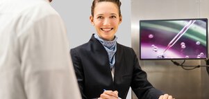 Eine junge Frau steht lächelnd an einer Art Empfang . Im Hintergrund sieht man auf einem Bildschirm eine kleine Pipette über einer Petrischale, die in Violettönen eingefärbt ist.