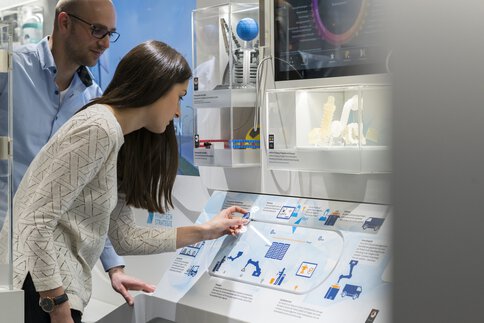 Eine Frau mit langen Haaren berührt eine interaktive Schautafel, während ein Mann mit Brille ihr über die Schulter sieht. Neben ihnen hängen weitere Schaukästen.