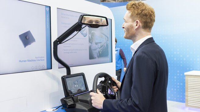 A man with red hair is using a driving simulator with on-screen displays and an intelligent rear-view mirror at an exhibition stand.
