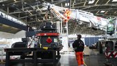 Ein Mann in Arbeitskleidung beobachtet, wie eine massive Industrieanlage mithilfe eines großen Autokrans innerhalb einer Messehalle angehoben wird.