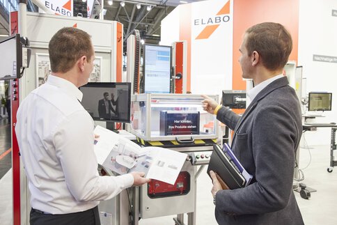 Zwei Männer betrachten an einem Stand ausgestellte Geräte, während sie Katalog und Notizbuch in der Hand halten.