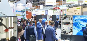 Zahlreiche Besucher, Messestände und Werbeschilder in der hell beleuchteten Messehalle A2.