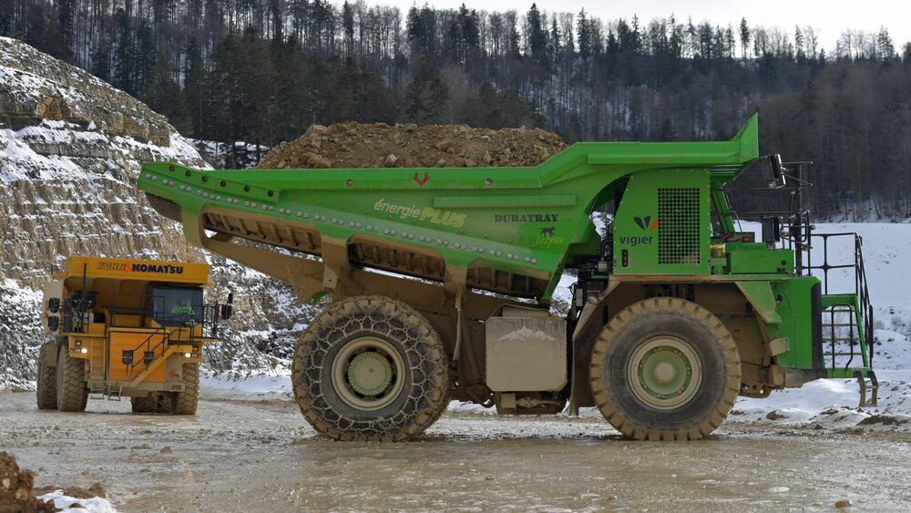 Zwei große Muldenkipper, gelb und grün, transportieren Erde in einem schneebedeckten Waldsteinbruch.