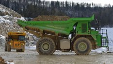 Two large dumper trucks, yellow and green, transport earth in a snow-covered forest quarry.
