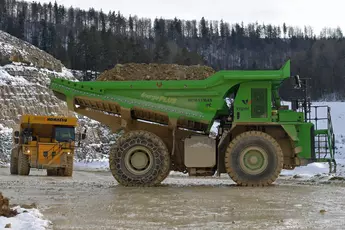 Zwei große Muldenkipper, gelb und grün, transportieren Erde in einem schneebedeckten Waldsteinbruch.