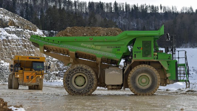Zwei große Muldenkipper, gelb und grün, transportieren Erde in einem schneebedeckten Waldsteinbruch.
