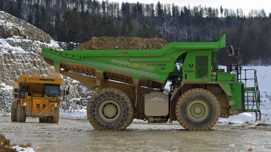 Two large dumper trucks, yellow and green, transport earth in a snow-covered forest quarry.
