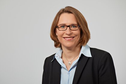 A smiling woman with glasses and short hair, wearing a black blazer and a light blue shirt.