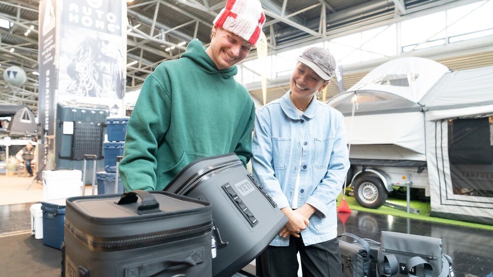 Zwei Aussteller präsentieren an einem Outdoor-Stand robuste Kühlboxen und Taschen vor einem ausgestellten Campinganhänger.