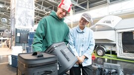 Zwei Aussteller präsentieren an einem Outdoor-Stand robuste Kühlboxen und Taschen vor einem ausgestellten Campinganhänger.