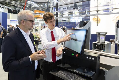 Zwei Männer stehen vor einem Computer an einem Ausstellerstand auf der productronica. Einer der Männer deutet auf den Bildschirm.