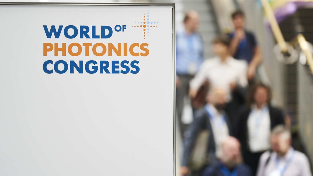 Sign with the logo of the World of Photonics Congress. Some people on an escalator can be seen blurred in the background.