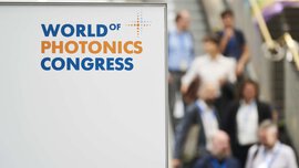 Sign with the logo of the World of Photonics Congress. Some people on an escalator can be seen blurred in the background.