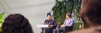 Two people are sitting on stage and conversing in front of an audience with green plants and a screen in the background.