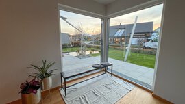 Blick aus dem Eckfenster auf einen modernen Garten, Bank und Teppich im Haus, Topfpflanzen auf Holzboden.