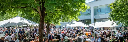 A crowd sits at tables between trees on a green area of the exhibition grounds with food and drinks.