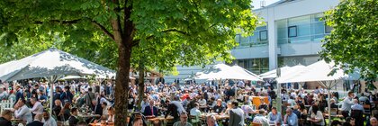 Eine Menschenmenge sitzt bei Essen und Getränken an Tischen zwischen Bäumen auf einer Grünfläche im Messegelände.