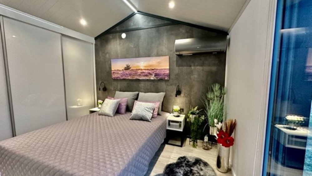 Modernes Schlafzimmer mit Doppelbett, grauer Steppdecke, Wandkunst und Pflanzen. Sanftes Licht schafft eine gemütliche Atmosphäre.