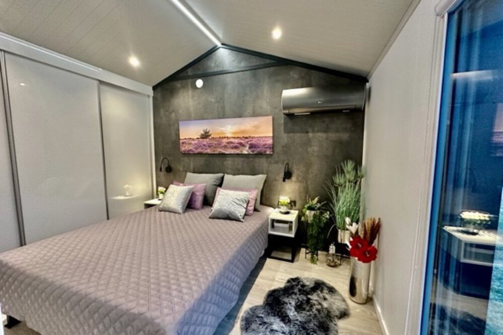 Modernes Schlafzimmer mit Doppelbett, grauer Steppdecke, Wandkunst und Pflanzen. Sanftes Licht schafft eine gemütliche Atmosphäre.