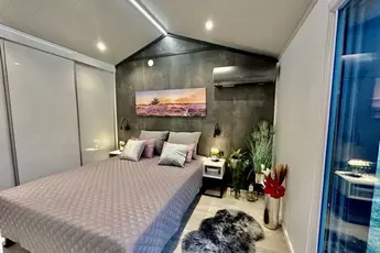 Modernes Schlafzimmer mit Doppelbett, grauer Steppdecke, Wandkunst und Pflanzen. Sanftes Licht schafft eine gemütliche Atmosphäre.