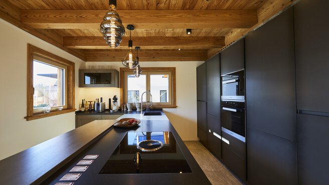 Moderne Küche mit dunklen Schränken, Holzdecke, eleganten Geräten und Hängelampen über einer zentralen Kochinsel.