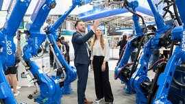 Zwei Personen stehen an einem Messestand und betrachten sechs blauen Roboterarmen, die rechts und links von ihnen Spalier stehen.