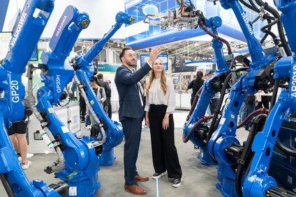 Two people are standing at an exhibition stand looking at six blue robotic arms lined up to their right and left.
