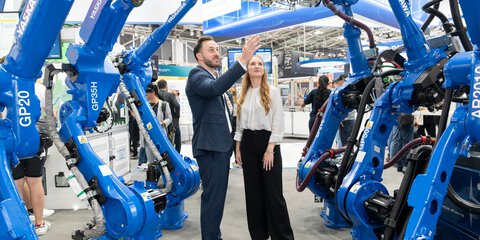 Two people are standing at an exhibition stand looking at six blue robotic arms lined up to their right and left.
