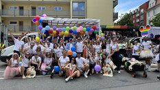 Eine Gruppe von Menschen in weißen Hemden mit Regenbogen-Accessoires posiert mit Luftballons bei einer Veranstaltung im Freien auf einer Stadtstraße.