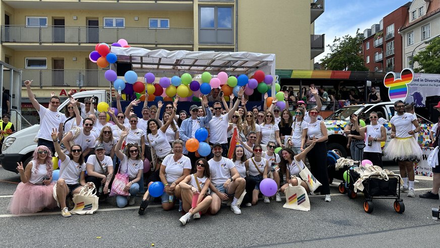Eine Gruppe von Menschen in weißen Hemden mit Regenbogen-Accessoires posiert mit Luftballons bei einer Veranstaltung im Freien auf einer Stadtstraße.