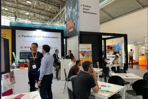 People networking and working at tables in an exhibition stand bearing the name “Pavilion Smart Maintenance+”.