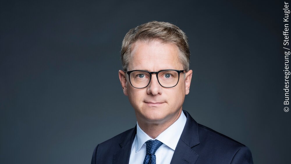 Ein Mann in einem dunklen Anzug, einem weißen Hemd und mit Brille posiert vor einem dunklen Hintergrund. Das Bild ist mit dem Vermerk © Bundesregierung/Steffen Kugler versehen.