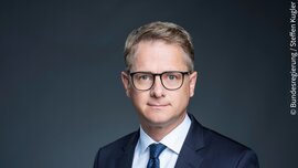 Ein Mann in einem dunklen Anzug, einem weißen Hemd und mit Brille posiert vor einem dunklen Hintergrund. Das Bild ist mit dem Vermerk © Bundesregierung/Steffen Kugler versehen.