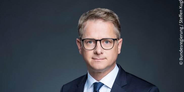 Ein Mann in einem dunklen Anzug, einem weißen Hemd und mit Brille posiert vor einem dunklen Hintergrund. Das Bild ist mit dem Vermerk © Bundesregierung/Steffen Kugler versehen.