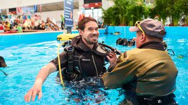 Ein Mann in Neoprenanzug und Taucherausrüstung steht in einem großen Pool in einer Messehalle, während ein anderer Mann ihm mit der Ausrüstung hilft.
