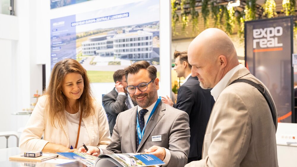 Three people in conversation at EXPO REAL 2026 in Munich, looking through a brochure together at an exhibition stand, with the EXPO REAL logo in the background.
