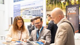 Three people in conversation at EXPO REAL 2026 in Munich, looking through a brochure together at an exhibition stand, with the EXPO REAL logo in the background.