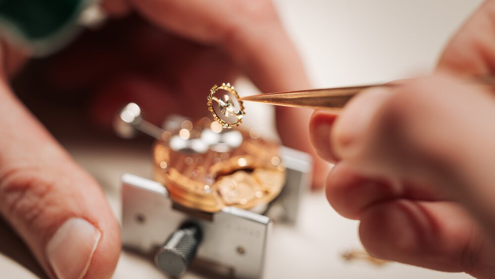 Eine Person benutzt eine Pinzette, um kleine Uhrenzahnräder mit einem Werkzeug zu reparieren, das die Uhr in Position hält.
