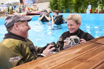 Ein Mann und eine Frau lehnen in einem Indoor-Schnorchel-Pool am Rand in Taucherkleidung und unterhalten sich.