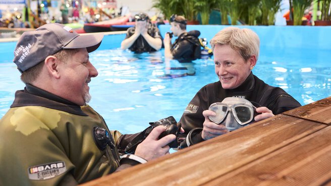 A man and a woman lean against the edge of an indoor snorkeling pool in diving gear and talk.