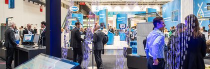 View of an exhibition hall with numerous visitors and stands through an exhibit with illuminated water droplets falling in patterns from a frame.