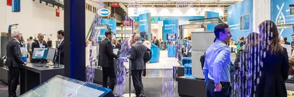 View of an exhibition hall with numerous visitors and stands through an exhibit with illuminated water droplets falling in patterns from a frame.