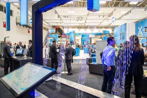 View of an exhibition hall with numerous visitors and stands through an exhibit with illuminated water droplets falling in patterns from a frame.