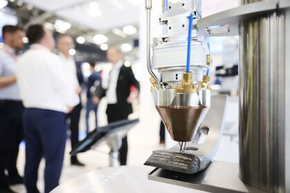 Close-up of an industrial laser head that precisely applies material to a metal surface during laser manufacturing; in the background, several people are conversing in a trade fair environment.