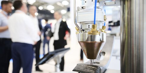 Close-up of an industrial laser head that precisely applies material to a metal surface during laser manufacturing; in the background, several people are conversing in a trade fair environment.