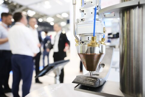 Nahaufnahme eines industriellen Laserkopfes, der bei der Laserfertigung präzise Material auf eine Metalloberfläche aufträgt; im Hintergrund unterhalten sich mehrere Personen in einer Messeumgebung.