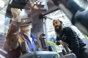Eine Person testet mit VR-Brille ein Baufahrzeug, während ein Experte daneben steht und das Szenario beobachtet.