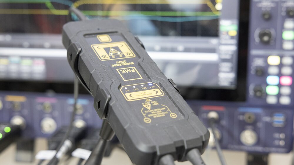 Close-up of the control unit of a PMK Bumblebee high-voltage differential probe in front of a blurred oscilloscope.