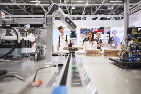 Menschen beobachten einen auf einem Messestand ausgestellten Roboterarm, der präzise Fräsungen an aufgestapelten Holzscheiben vornimmt.